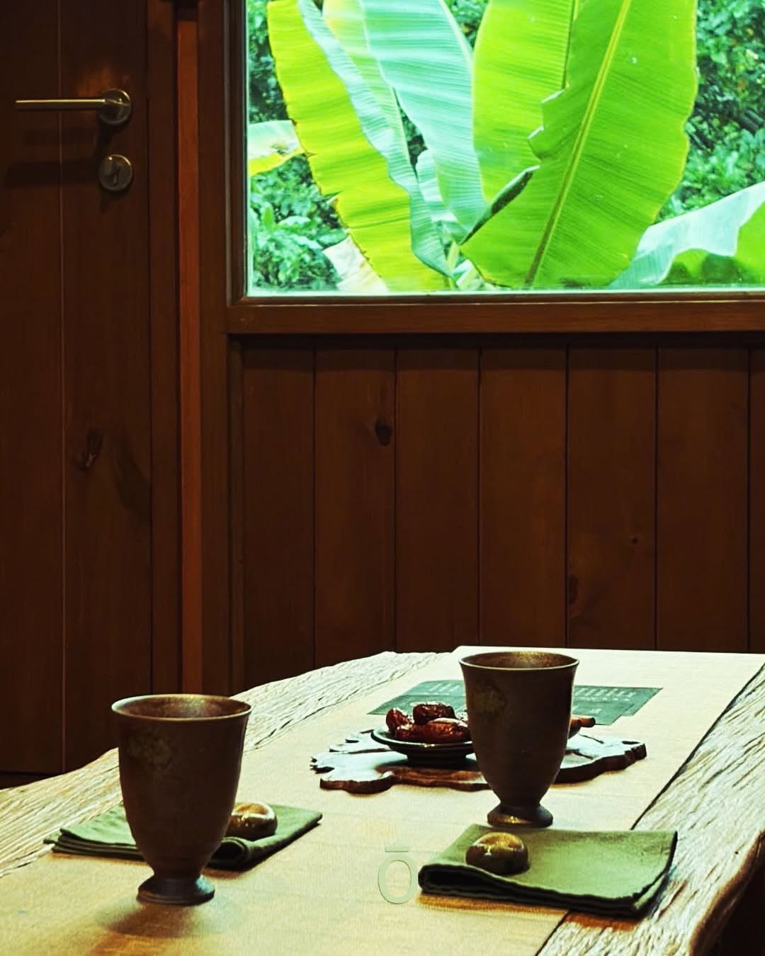 A wooden table with two ceramic cups and a plate of dates, by a window looking out at green banana leaves