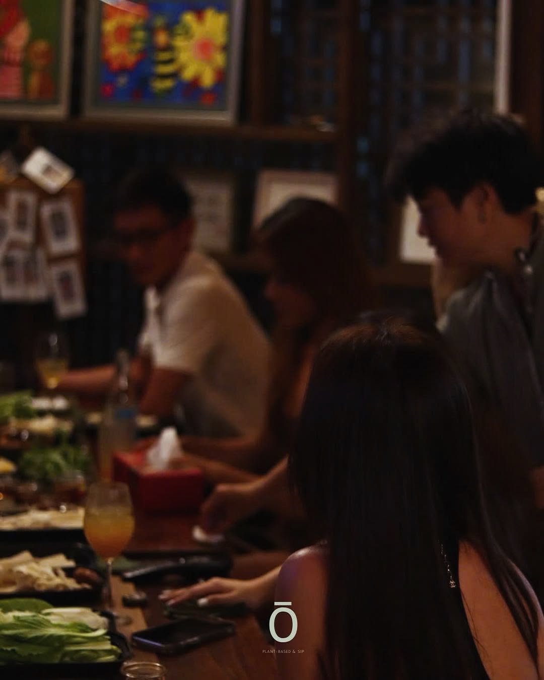 Guests around a long table at dinner, glasses catching the warm light, fresh greens on the table