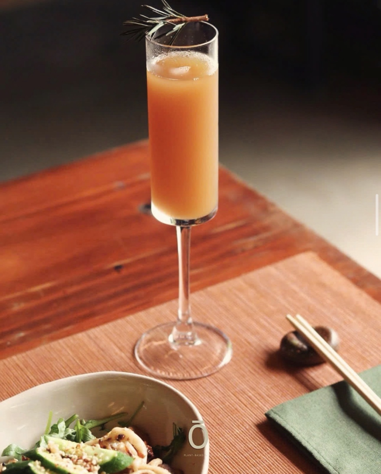 A pale orange cocktail topped with a rosemary sprig on a wooden table, beside a salad plate and chopsticks