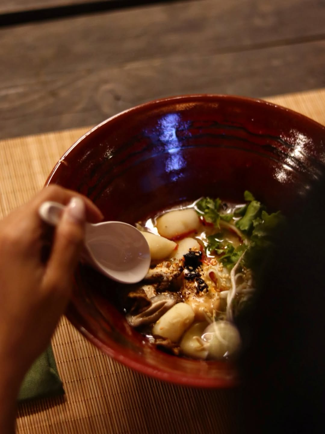 A hand holding a ceramic spoon over a brown clay bowl of mushroom soup with gnocchi and fresh greens, on a wooden table